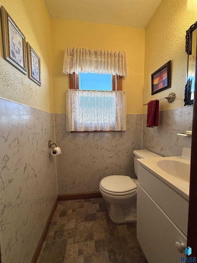 half bathroom with baseboards, toilet, a wainscoted wall, stone finish flooring, and vanity