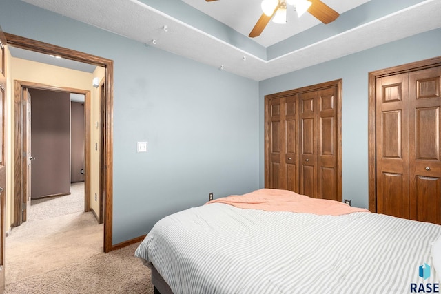 bedroom featuring light carpet, baseboards, ceiling fan, a tray ceiling, and multiple closets