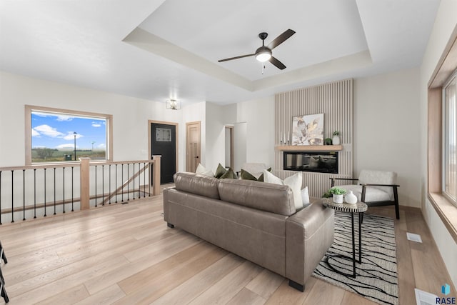 living area with a tray ceiling, visible vents, a ceiling fan, a glass covered fireplace, and wood finished floors