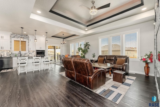 living room featuring dark wood-type flooring, a raised ceiling, plenty of natural light, and ceiling fan with notable chandelier