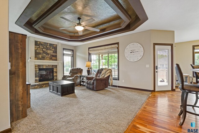 living area featuring a ceiling fan, a tray ceiling, wood finished floors, a stone fireplace, and baseboards