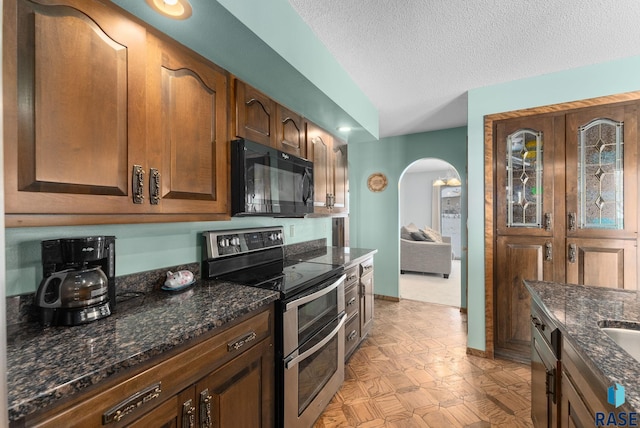 kitchen with a textured ceiling, dark stone counters, arched walkways, black microwave, and range with two ovens