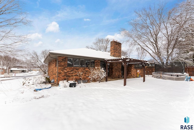 snow covered back of property featuring brick siding, a trampoline, fence, a chimney, and a pergola