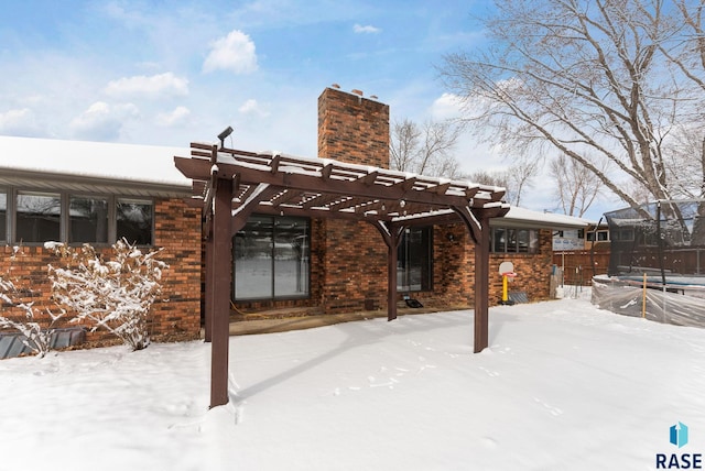 snow covered rear of property with brick siding, a chimney, a trampoline, and a pergola