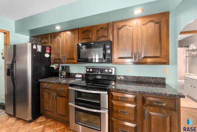 kitchen with a textured ceiling, dark stone countertops, brown cabinets, and stainless steel appliances