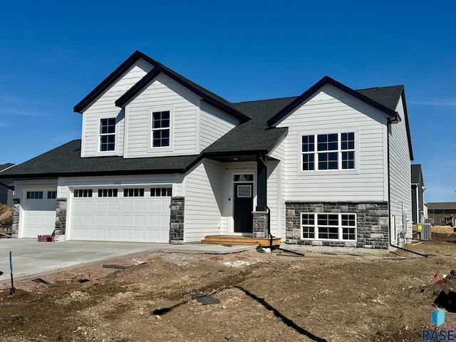 view of front facade featuring central air condition unit, roof with shingles, a garage, stone siding, and driveway