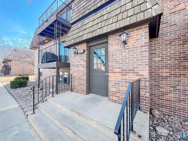 view of exterior entry featuring brick siding, mansard roof, and a balcony