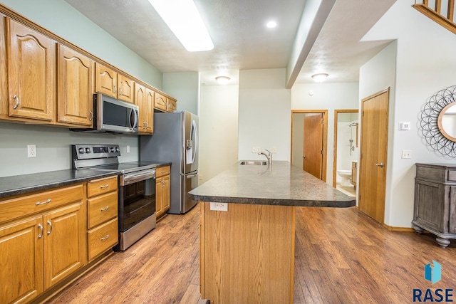 kitchen with light wood-type flooring, a center island with sink, a sink, dark countertops, and appliances with stainless steel finishes