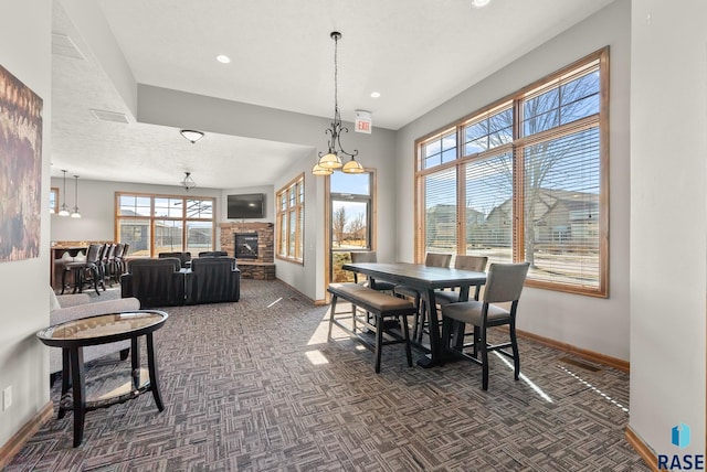 dining room featuring visible vents, baseboards, a stone fireplace, a notable chandelier, and dark carpet