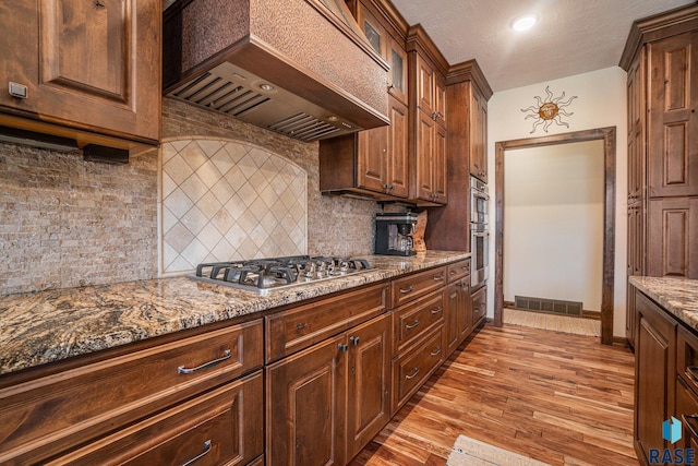 kitchen with light stone countertops, visible vents, premium range hood, decorative backsplash, and light wood-type flooring