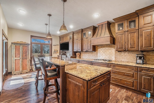 kitchen featuring a center island with sink, backsplash, custom exhaust hood, and dark wood finished floors