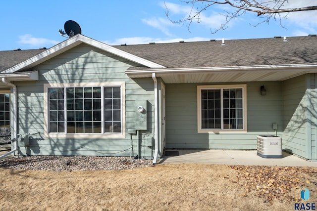 view of exterior entry featuring central AC unit, a shingled roof, and a patio