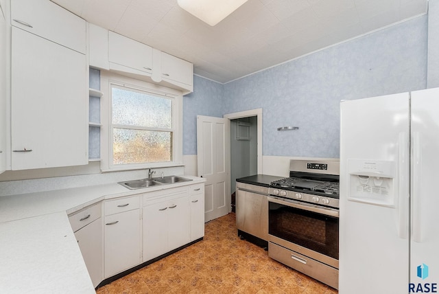 kitchen featuring wallpapered walls, stainless steel range with gas stovetop, a sink, white cabinets, and white fridge with ice dispenser