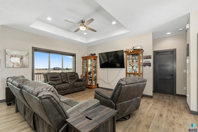living room with light wood-type flooring, a tray ceiling, baseboards, and ceiling fan