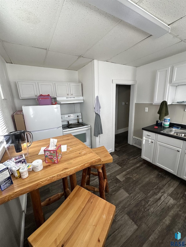 kitchen featuring white appliances, under cabinet range hood, a drop ceiling, dark wood-style floors, and white cabinets