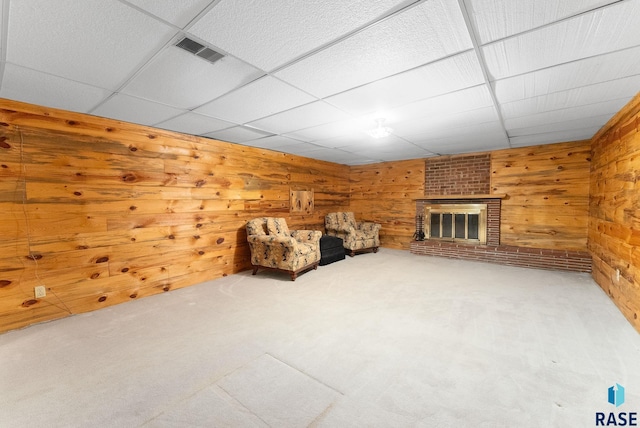 unfurnished room with visible vents, wood walls, a brick fireplace, a drop ceiling, and carpet floors