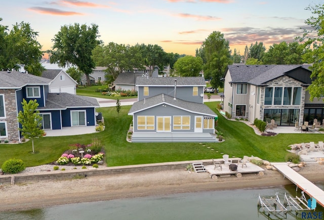 back of property at dusk featuring roof with shingles, a patio, a residential view, a water view, and a yard