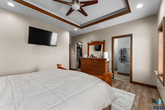 bedroom featuring a spacious closet, recessed lighting, a raised ceiling, wood-type flooring, and baseboards
