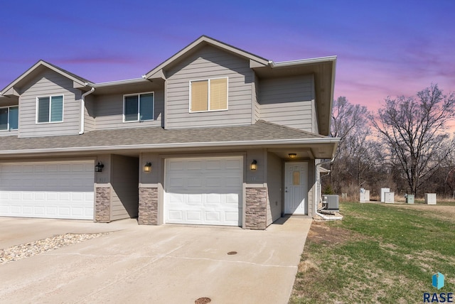 view of property with central air condition unit, stone siding, an attached garage, a shingled roof, and concrete driveway