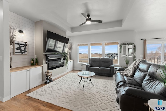 living area featuring a ceiling fan, a raised ceiling, a glass covered fireplace, and a healthy amount of sunlight