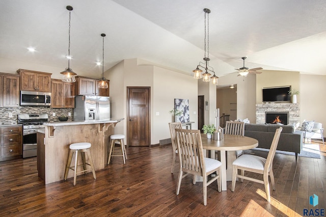 dining space featuring dark wood finished floors, a stone fireplace, a ceiling fan, and vaulted ceiling