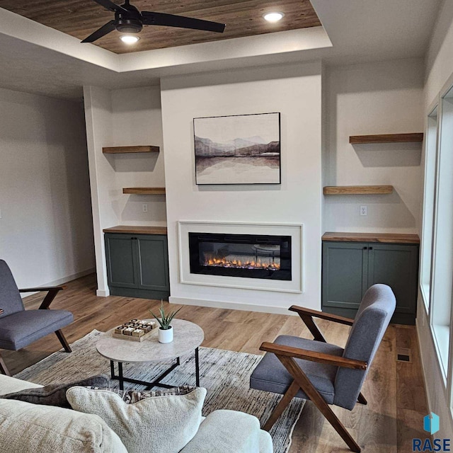 living area featuring a glass covered fireplace, a tray ceiling, wood finished floors, and ceiling fan