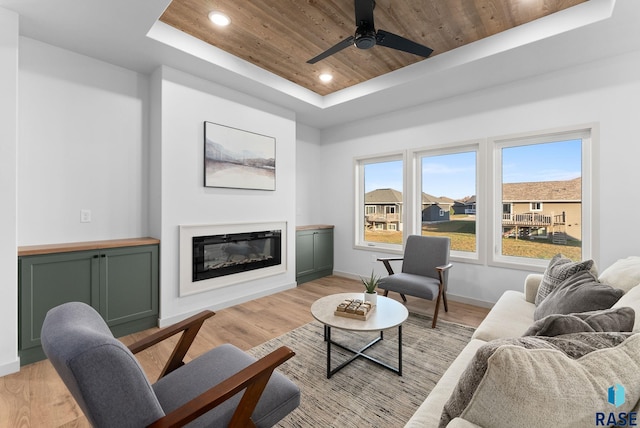 living area featuring wooden ceiling, ceiling fan, a tray ceiling, light wood-style flooring, and a glass covered fireplace