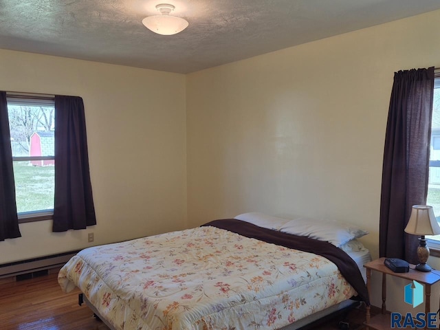 bedroom featuring a textured ceiling, wood finished floors, a baseboard heating unit, and multiple windows