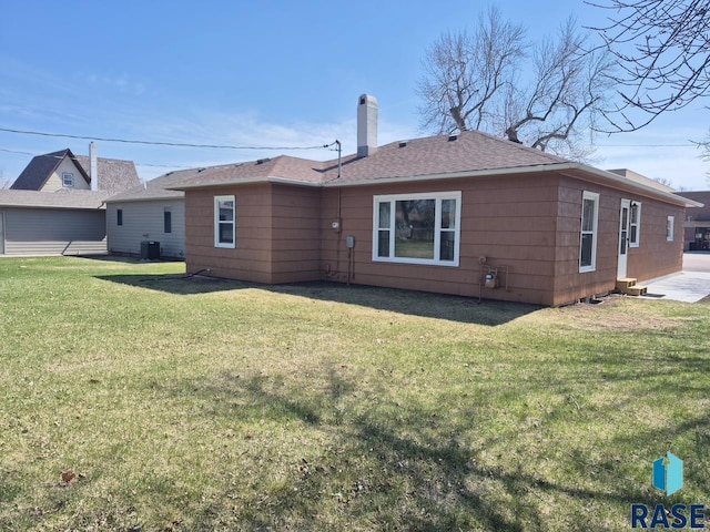 rear view of house with a yard, a shingled roof, central AC, and a chimney