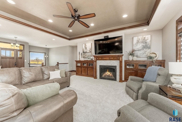 carpeted living room with ceiling fan with notable chandelier, a tiled fireplace, a raised ceiling, ornamental molding, and recessed lighting