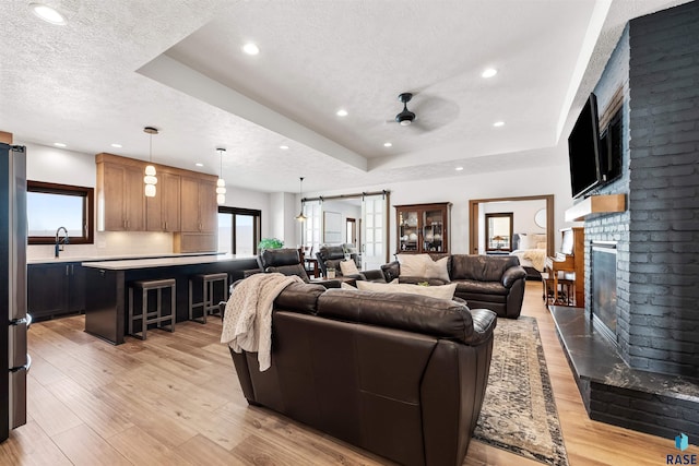 living room featuring a ceiling fan, a textured ceiling, a tray ceiling, light wood finished floors, and a brick fireplace