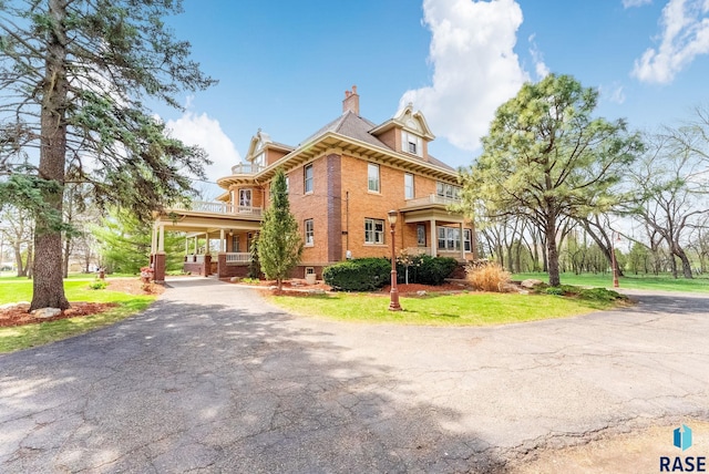 victorian-style house featuring a chimney, brick siding, a balcony, and driveway