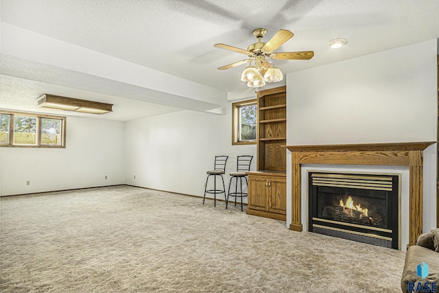 unfurnished living room featuring a warm lit fireplace, ceiling fan, a textured ceiling, carpet, and baseboards