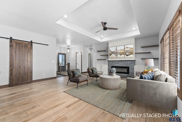 living room with a ceiling fan, a stone fireplace, a barn door, wood finished floors, and a raised ceiling