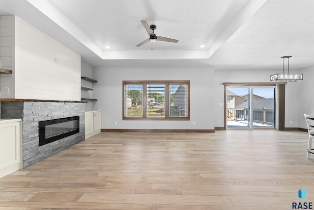 unfurnished living room featuring a fireplace, baseboards, ceiling fan with notable chandelier, a tray ceiling, and light wood-style flooring