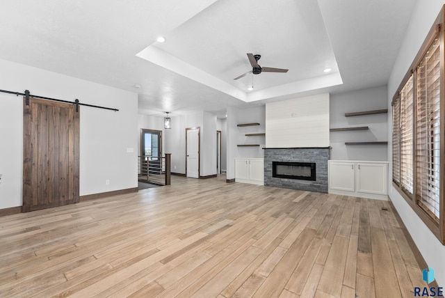 unfurnished living room featuring ceiling fan, a barn door, light wood finished floors, a fireplace, and a tray ceiling
