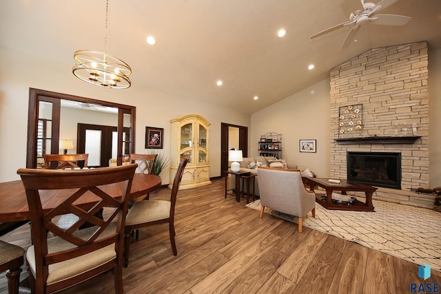 dining space featuring wood finished floors, a stone fireplace, high vaulted ceiling, recessed lighting, and ceiling fan with notable chandelier