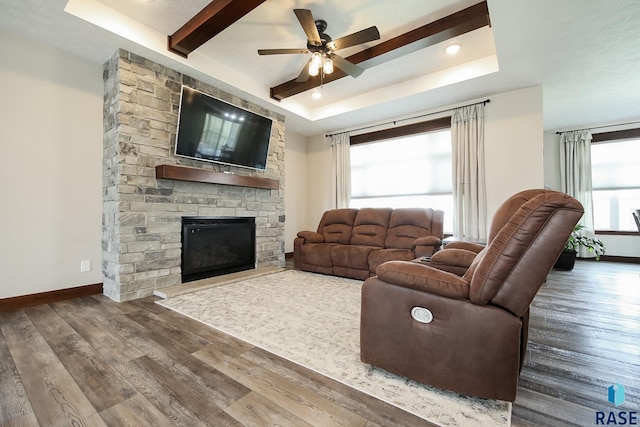 living area with a ceiling fan, a stone fireplace, wood finished floors, beam ceiling, and a tray ceiling