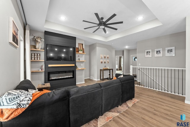 living area with a tray ceiling, a large fireplace, a ceiling fan, and light wood-style flooring