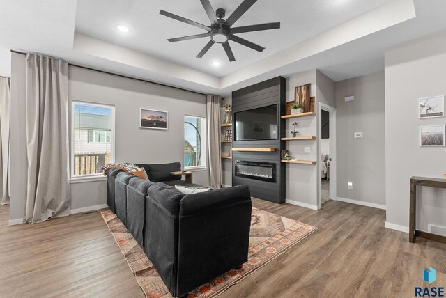 living room with a raised ceiling, a glass covered fireplace, light wood-style flooring, a ceiling fan, and baseboards