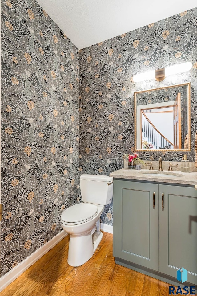 half bathroom with wood finished floors, vanity, baseboards, toilet, and wallpapered walls