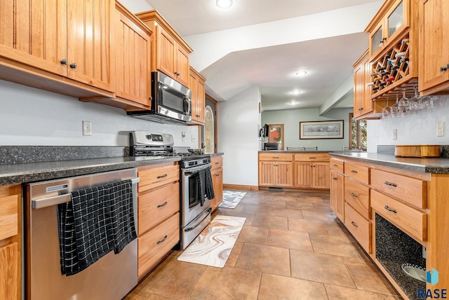 kitchen with dark countertops, appliances with stainless steel finishes, glass insert cabinets, and light brown cabinets