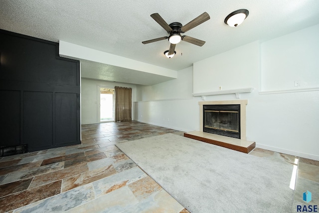 unfurnished living room featuring stone tile floors, ceiling fan, a glass covered fireplace, and a textured ceiling