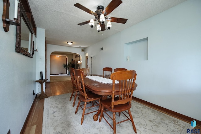 dining room featuring arched walkways, a ceiling fan, light wood-type flooring, a textured ceiling, and baseboards
