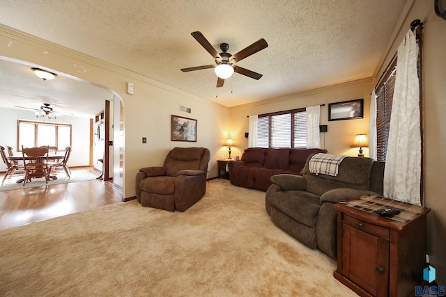 living area featuring ceiling fan, arched walkways, ornamental molding, light colored carpet, and a textured ceiling