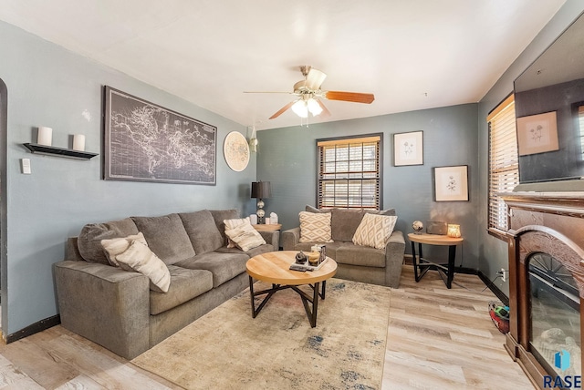 living area featuring light wood-type flooring, a ceiling fan, a glass covered fireplace, and baseboards