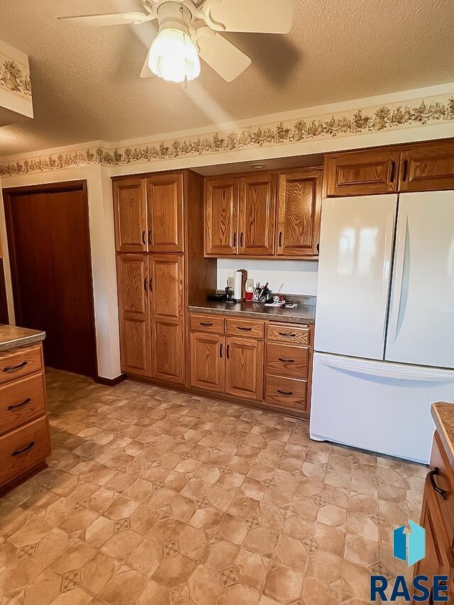 515 151st St, Luverne, MN 56156 - kitchen with freestanding refrigerator, brown cabinetry, a textured ceiling, ceiling fan, and dark countertops