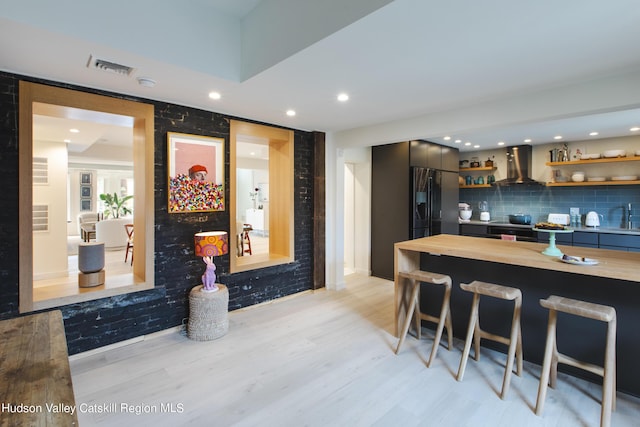 kitchen with fridge with ice dispenser, wall chimney range hood, light hardwood / wood-style flooring, butcher block countertops, and a kitchen bar