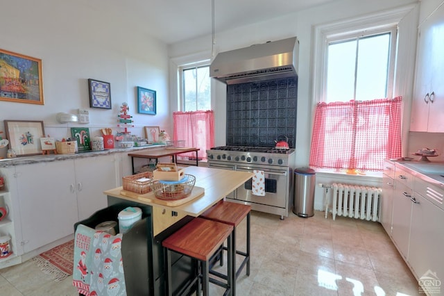kitchen with range with two ovens, wall chimney range hood, radiator heating unit, white cabinetry, and light countertops
