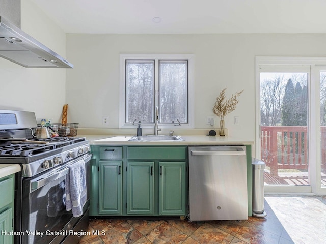 kitchen with stainless steel appliances, a sink, light countertops, wall chimney range hood, and green cabinetry